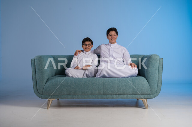 الجلوس والنظر إلى الكاميرا, قضاء أوقات ممتعة مع الأخوات, تعزيز وتقوية العلاقة الإيجابية الأسرية, مفهوم الحب والحنان بين الأخوة, بورتريه لأخيين عربيين خليجين سعوديين يرتديان الثوب التقليدي يجلسان على كنبه مريحة, خلفية زرقاء