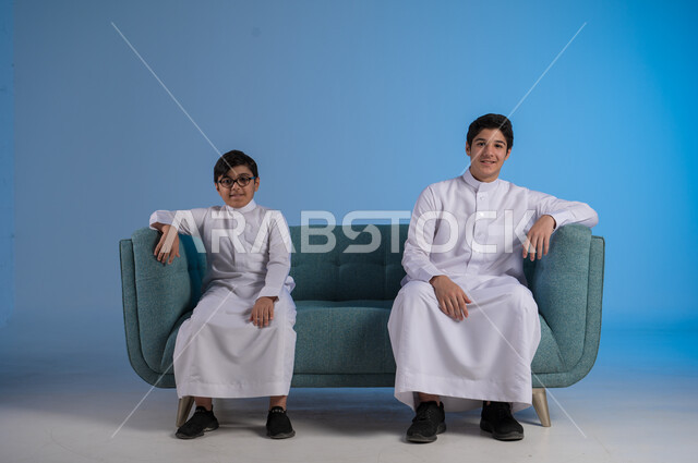 الجلوس والنظر إلى الكاميرا, التسلية وقضاء الوقت الممتع مع الأخوات, تقوية الروابط الاسرية, بورتريه لشاب عربي خليجي سعودي يرتدي الثوب التقليدي يضع يده على كتف أخيه, لحظات مليئة بالحنان والحب,  خلفية زرقاء