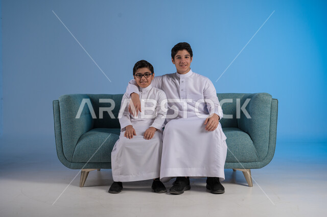 التسلية وقضاء الوقت الممتع مع الأخوات, تقوية الروابط الاسرية, بورتريه لشاب عربي خليجي سعودي يرتدي الثوب التقليدي يضع يده على كتف أخيه, لحظات مليئة بالحنان والحب, الجلوس والنظر إلى الكاميرا, خلفية زرقاء