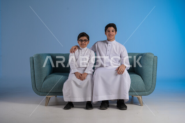 التسلية وقضاء الوقت الممتع مع الأخوات, تقوية الروابط الاسرية, بورتريه لشاب عربي خليجي سعودي يرتدي الثوب التقليدي يضع يده على كتف أخيه, لحظات مليئة بالحنان والحب, الجلوس والنظر إلى الكاميرا, خلفية زرقاء