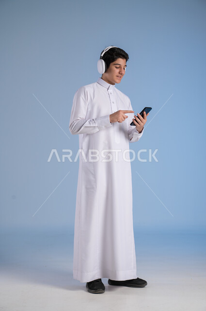 التقنيات والتكنولوجيا الحديثة، بورتريه لشاب عربي سعودي خليجي يرتدي الثوب التقليدي يضع سماعات الهيدفون، التواصل مع الاهل والاقارب عبر الهاتف المحمول، مفهوم الاستماع والاصغاء، الانسجام في سماع الموسيقا والتسجيلات الصوتية، الجسم بالطول الكامل، خلفية زرقاء