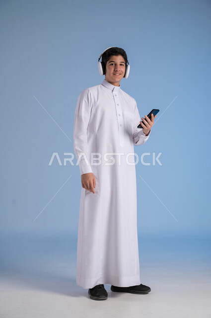 التقنيات والتكنولوجيا الحديثة، بورتريه لشاب عربي سعودي خليجي يرتدي الثوب التقليدي يضع سماعات الهيدفون، التواصل مع الاهل والاقارب عبر الهاتف المحمول، مفهوم الاستماع والاصغاء، الانسجام في سماع الموسيقا والتسجيلات الصوتية، الجسم بالطول الكامل، خلفية زرقاء
