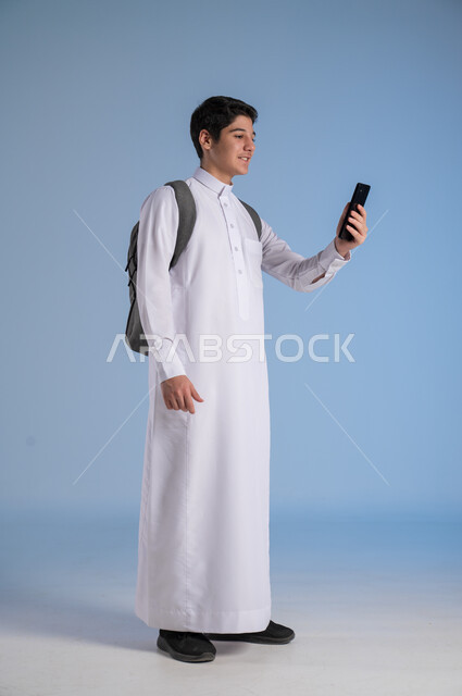 The concept of distance education, the development of learning methods in Saudi Arabia, the use of modern technical devices, online communication, a portrait of a Saudi Arabian Gulf student wearing traditional dress and a school backpack holding a mobile phone in his hand, a full-length body image, blue background