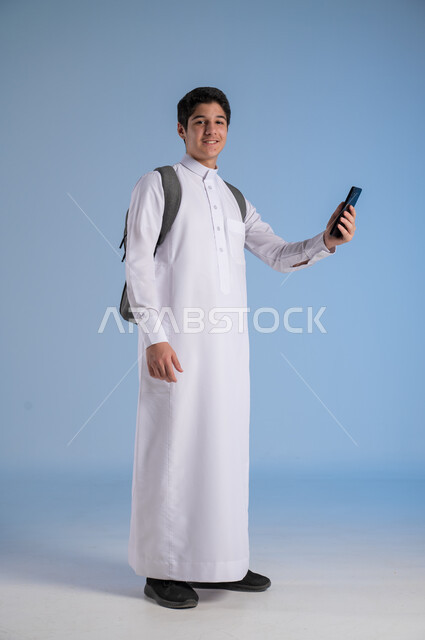 Development of learning methods in Saudi Arabia, use of modern technology devices, online communication, portrait of a Saudi Arabian Gulf student wearing traditional dress and school backpack holding a mobile phone, concept of distance education, full-length body image, blue background