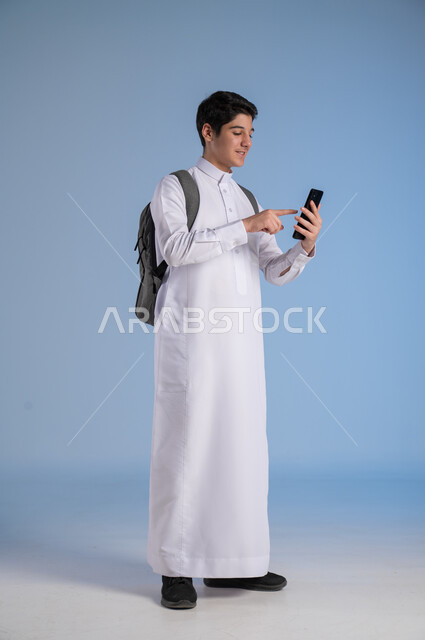 Development of learning methods in Saudi Arabia, use of modern technology devices, online communication, portrait of a Saudi Arabian Gulf student wearing traditional dress and school backpack holding a mobile phone, concept of distance education, full-length body image, blue background