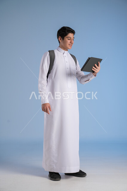 Using modern and advanced devices and technologies, the concept of distance learning, a portrait of a Saudi Arabian Gulf boy wearing traditional clothing, putting on a school backpack and holding a tablet in his hand, following classes online, a full body image, blue background