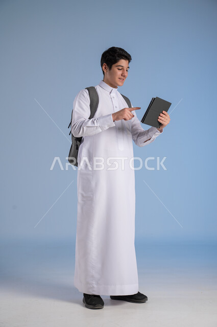التفاعل مع الحصص الدرسية عبر الأونلاين, مفهوم التعليم عن بعد, بورتريه لولد عربي خليجي سعودي يرتدي الثوب التقليدي وحقيبة الظهر المدرسية يحمل بيده جهاز التابليت, صورة الجسم بالطول الكامل, خلفية زرقاء