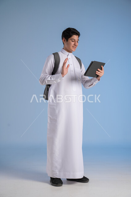 اجراء مكالمة فيديو عبر الأجهزة والتقنيات الحديثة والمتطورة, مفهوم التعلم عن بعد, بورتريه لولد عربي خليجي سعودي يرتدي الثوب التقليدي يضع حقيبة الظهر المدرسية ويحمل بيده جهاز التابليت, متابعة الحصص عبر الأونلاين, صورة الجسم بالكامل, خلفية زرقاء