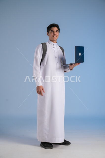 مفهوم التعلم عن بعد, متابعة الحصص عبر الأونلاين, تطور وسائل التعليم في المملكة, بورتريه لولد عربي خليجي سعودي يرتدي الثوب التقليدي يضع حقيبة الظهر المدرسية ويحمل بيده جهاز الحاسوب, صورة الجسم بالطول الكامل, خلفية زرقاء