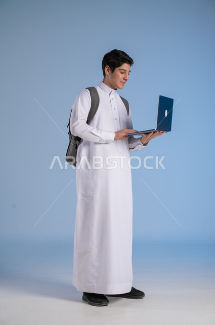  تطور وسائل التعليم في المملكة, مفهوم التعلم عن بعد, متابعة الحصص عبر الأونلاين, بورتريه لولد عربي خليجي سعودي يرتدي الثوب التقليدي يضع حقيبة الظهر المدرسية ويحمل بيده جهاز الحاسوب, صورة الجسم بالطول الكامل, خلفية زرقاء