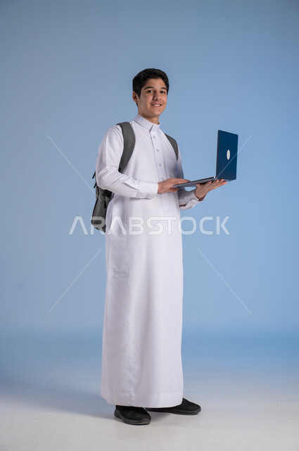 The concept of distance learning, following classes online, the development of educational methods in the Kingdom, a portrait of a Saudi Arabian Gulf boy wearing traditional clothing, putting on a school backpack and holding a computer in his hand, a full-length body image, blue background