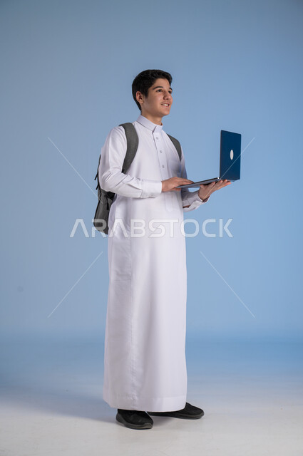 Looking at something, distance learning concept, following classes online, development of education methods in the Kingdom, portrait of a Saudi Arabian Gulf boy wearing traditional dress, putting on a school backpack and holding a computer in his hand, full-length body image, blue background
