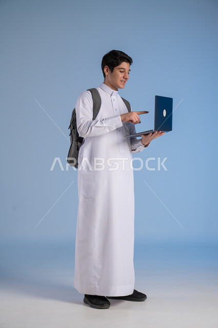 مفهوم التعلم عن بعد, متابعة الحصص عبر الأونلاين, تطور وسائل التعليم في المملكة, بورتريه لولد عربي خليجي سعودي يرتدي الثوب التقليدي يضع حقيبة الظهر المدرسية ويحمل بيده جهاز الحاسوب, صورة الجسم بالطول الكامل, خلفية زرقاء