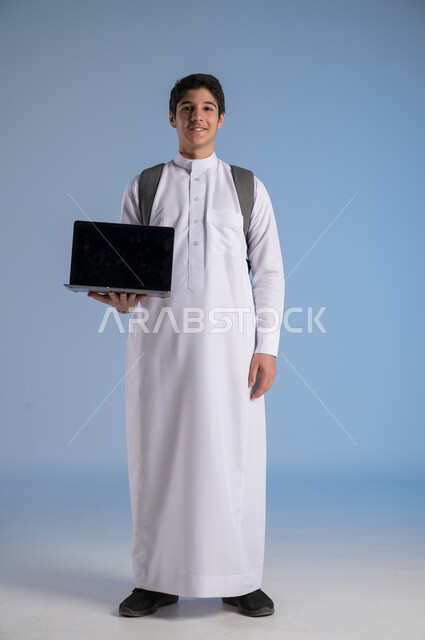 استخدام الأجهزة والتقنيات الحديثة, مفهوم التعليم عبر الانترنت, متابعة الحصص عبر الأونلاين, بورتريه لولد عربي خليجي سعودي يرتدي الثوب التقليدي يضع حقيبة الظهر المدرسية ويمسك بيده الابتوب, عرض شاشة سوداء فارغة على الحاسوب, صورة الجسم بالكامل, خلفية زرقاء
