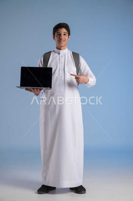 عرض شاشة سوداء فارغة على الحاسوب, استخدام الأجهزة والتقنيات الحديثة, مفهوم التعليم عبر الانترنت, متابعة الحصص عبر الأونلاين, بورتريه لولد عربي خليجي سعودي يرتدي الثوب التقليدي يضع حقيبة الظهر المدرسية ويمسك بيده الابتوب, صورة الجسم بالكامل, خلفية زرقاء