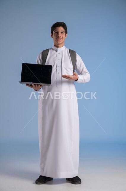 Blank black screen on computer, use of modern devices and technologies, concept of online education, follow-up of classes online, portrait of a Saudi Arabian Gulf boy wearing traditional dress, putting on a school backpack and holding a laptop in his hand, full body image, blue background