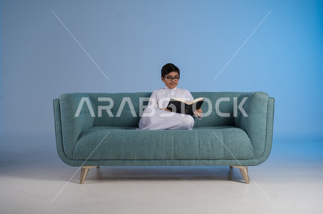 حب المطالعة والقراءة, قضاء أوقات الفراغ بالتثقيف والتعلم, تنمية مهارات القراءة, الاهتمام بتصفح الكتب المفيدة, بورتريه لفتى عربي خليجي سعودي يرتدي الثوب التقليدي والنظارة الطبية يجلس على كنبه مريحة ويمسك بيده الكتاب, خلفية رزقاء