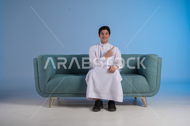 الاشارة إلى شيء ما وتحديد مكان معين, بورتريه لشاب عربي خليجي سعودي يرتدي الثوب التقليدي يجلس على كنبه مريحة, النظر إلى الكاميرا بإيماءات السرور, خلفية رزقاء