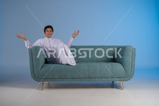 عدم المعرفة والشعور بالحيرة, الجلوس في غرفة المعيشة ورفع اليدين للأعلى, بورتريه لشاب عربي خليجي سعودي يرتدي الثوب التقليدي يجلس على كنبه مريحة, النظر إلى الكاميرا بإيماءات السرور, خلفية رزقاء