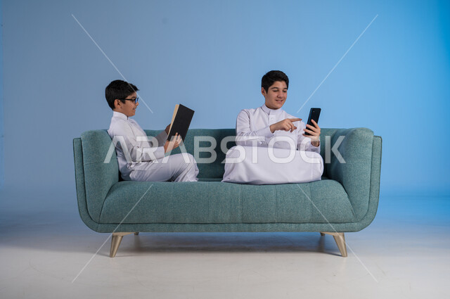 التعاون بين الأخوة لإنجاز المهام عن بعد, تصفح مواقع التواصل الاجتماعي, بورتريه لأخيين عربيين خليجيين سعوديين يرتديان الثوب التقليدي يستخدمان الأجهزة التقنية الحديثة, التواصل مع الأصدقاء عبر الأونلاين,  خلفية زرقاء