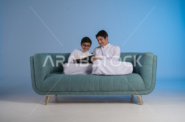 التعاون بين الأخوة لإنجاز المهام عن بعد, تصفح مواقع التواصل الاجتماعي, بورتريه لأخيين عربيين خليجيين سعوديين يرتديان الثوب التقليدي يستخدمان الأجهزة التقنية الحديثة, التواصل مع الأصدقاء عبر الأونلاين,  خلفية زرقاء