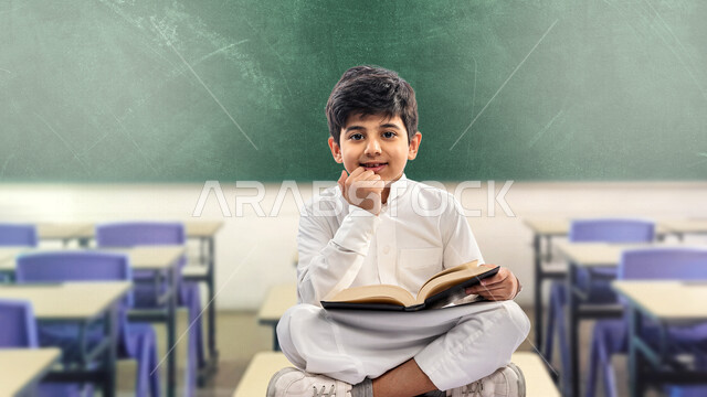 صورة لتلميذ مدرسي عربي خليجي سعودي مبتسم ، يجلس في غرفة صفية ، يرتدي الثوب السعودي التقليدي ، بيده كتاب مفتوح يقرأ به ، ايماءات و تعابير وجه و يدين مختلفة ، الاستمتاع بقراءة الكتاب المدرسي ، مدرسة سعودية