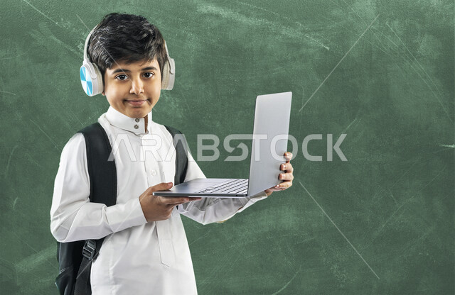 بورتريه لتلميذ مدرسي عربي خليجي سعودي مبتسم ، يرتدي حقيبة ظهر مدرسية  و سماعة الرأس ، يرتدي الثوب السعودي التقليدي ، بيده حاسوب محمول ، ايماءات و تعابير وجه و يدين مختلفة ، الدراسة من خلال منصة تعليمية للتعلم عن بعد ،  التعلم عبر الانترنت