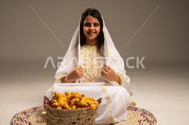 A basket full of fresh dates, national agricultural crops, a close-up portrait of a smiling Saudi Arabian Gulf girl wearing a shayla and a neshel sitting on a straw rug, looking at the camera with gestures of pride and honor, the day we started 1727 AD, wearing the costumes of the Eastern Province, the anniversary of the founding of the first Saudi state, February 22, beige background