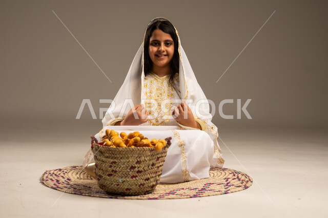 A basket full of fresh dates, national agricultural crops, a close-up portrait of a smiling Saudi Arabian Gulf girl wearing a shayla and a neshel sitting on a straw rug, looking at the camera with gestures of pride and honor, the day we started 1727 AD, wearing the costumes of the Eastern Province, the anniversary of the founding of the first Saudi state, February 22, beige background