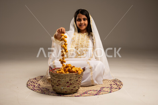 النظر الى الكاميرا بتعابير الفخر والاعتزاز، بورتريه مقرب  لفتاة عربية خليجية سعودية ترتدي الشيلة والنشل تمسك بيدها حبات البلح الطازج، يوم بدينا١٧٢٧م، ارتداء أزياء المنطقة الشرقية، ذكرى تأسيس الدولة السعودية الأولى٢٢فبراير،  منتجات ومحاصيل زراعية محلية وطنية، خلفية بيج