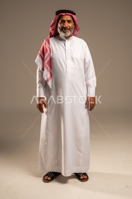 مفهوم الهيبة والوقار، تمثيل الرجولة العربية الأصيلة، بورتريه لرجل عربي خليجي سعودي كبير السن يرتدي الشماغ والثوب التقليدي ينظر الى الكاميرا بايماءات الفرح السرور، الاهتمام بالأناقة والمظهر الخارجي، صوة الجسم بالطول الكامل, خلفية بيج