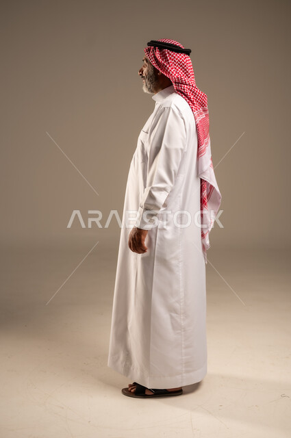 الاهتمام بالأناقة والمظهر الخارجي، مفهوم الهيبة والوقار، تمثيل الرجولة العربية الأصيلة، بورتريه من الجانب لرجل عربي خليجي سعودي كبير السن يرتدي الشماغ والثوب التقليدي يقف باستقامة ينظر الى شيء ما بايماءات الفرح السرور، صوة الجسم بالطول الكامل, خلفية بيج