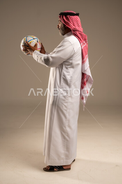 الحماس والتفاعل مع المنتخب، إيماءات الشغف والتشجيع للفريق، بورتريه من الجانب لرجل كبير السن عربي خليجي سعودي مبتسم يرتدي الشماغ والثوب التقليدي يحمل بيده كرة القدم، أنديه رياضية مختلفة، صوة الجسم بالطول الكامل, خلفية بيج