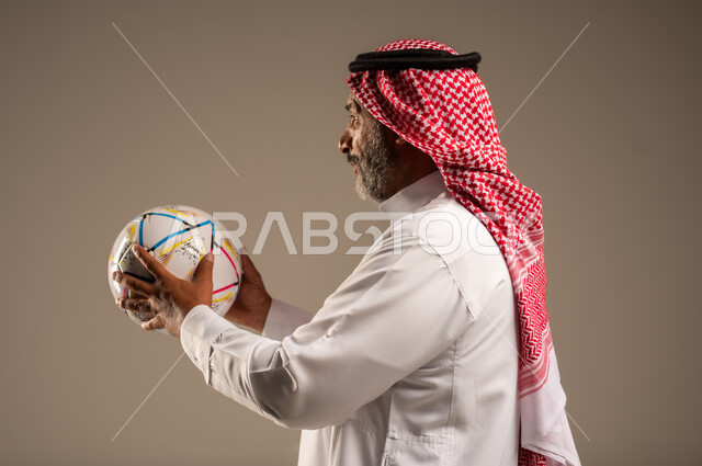 الحماس والتفاعل مع المنتخب، إيماءات الشغف والتشجيع للفريق، بورتريه مقرب من الجانب لرجل كبير السن عربي خليجي سعودي مبتسم يرتدي الشماغ والثوب التقليدي يحمل بيده كرة القدم، أنديه رياضية مختلفة، خلفية بيج