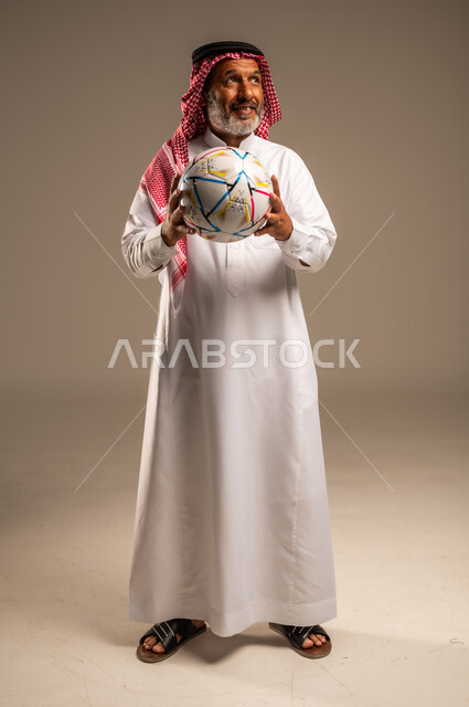 الحماس والتفاعل مع المنتخب، إيماءات الشغف والتشجيع للفريق، بورتريه لرجل كبير السن عربي خليجي سعودي مبتسم يرتدي الشماغ والثوب التقليدي ينظر الى شيء ما يحمل بيده كرة القدم، أنديه رياضية مختلفة، صوة الجسم بالطول الكامل, خلفية بيج