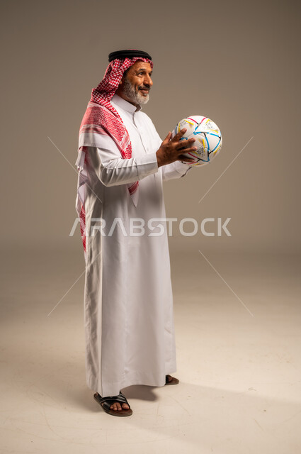 الحماس والتفاعل مع المنتخب، إيماءات الشغف والتشجيع للفريق، بورتريه لرجل كبير السن عربي خليجي سعودي مبتسم يرتدي الشماغ والثوب التقليدي ينظر الى شيء ما يحمل بيده كرة القدم، أنديه رياضية مختلفة، صوة الجسم بالطول الكامل, خلفية بيج