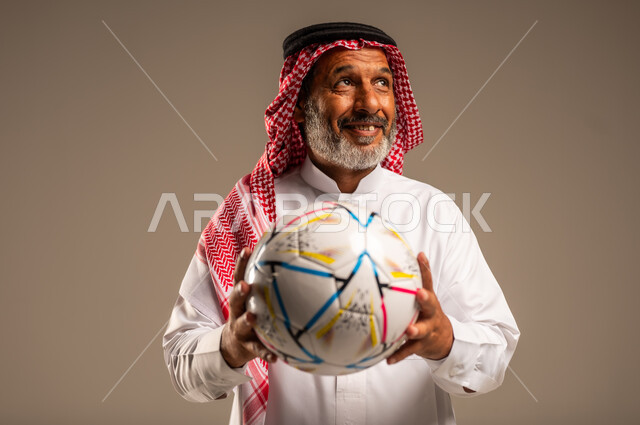 أنديه رياضية مختلفة، الحماس والتفاعل مع المنتخب، إيماءات الشغف والتشجيع للفريق، بورتريه مقرب لرجل كبير السن عربي خليجي سعودي مبتسم يرتدي الشماغ والثوب التقليدي ينظر الى شيء ما يحمل بيده كرة القدم، خلفية بيج