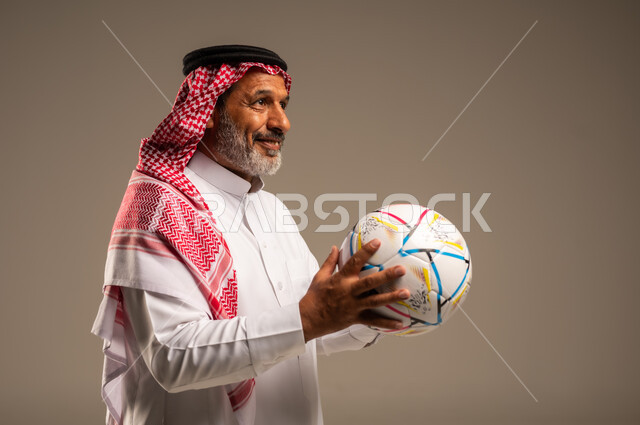 أنديه رياضية مختلفة، الحماس والتفاعل مع المنتخب، إيماءات الشغف والتشجيع للفريق، بورتريه مقرب لرجل كبير السن عربي خليجي سعودي مبتسم يرتدي الشماغ والثوب التقليدي ينظر الى شيء ما يحمل بيده كرة القدم، خلفية بيج