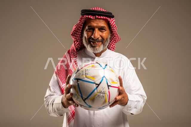 أنديه رياضية مختلفة، الحماس والتفاعل مع المنتخب، إيماءات الشغف والتشجيع للفريق، بورتريه مقرب لرجل كبير السن عربي خليجي سعودي مبتسم يرتدي الشماغ والثوب التقليدي ينظر الى الكاميرا بتعابير السرور يحمل بيده كرة القدم، خلفية بيج