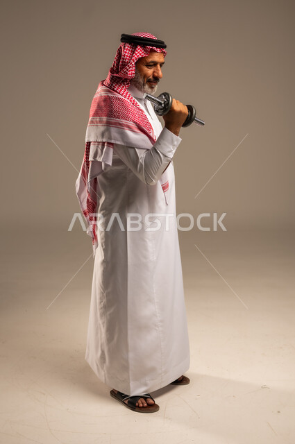 تمارين رفع الأثقال (دمبلز)، ممارسة التمارين الرياضية مع الدمبل، بورتريه من الجانب لرجل كبير السن عربي خليجي سعودي مبتسم يرتدي الشماغ والثوب التقليدي يقوم برفع الأوزان الحديدية الثقيلة، المحافظة على الصحة واللياقة الجسدية، صورة الجسم بالطول الكامل, خلفية بيج