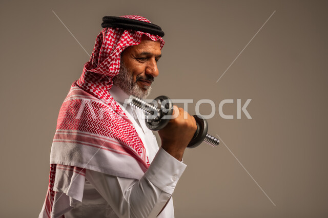 تمارين رفع الأثقال (دمبلز)، ممارسة التمارين الرياضية مع الدمبل، بورتريه مقرب من الجانب لرجل كبير السن عربي خليجي سعودي مبتسم يرتدي الشماغ والثوب التقليدي يقوم برفع الأوزان الحديدية الثقيلة، المحافظة على الصحة واللياقة الجسدية، خلفية بيج