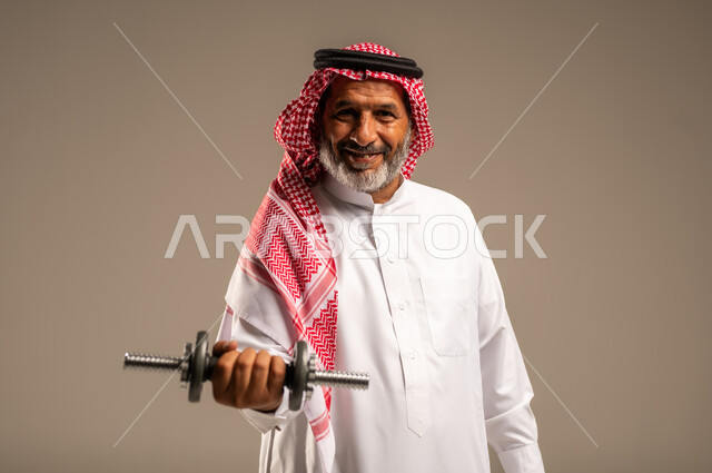 تمارين رفع الأثقال (دمبلز)، ممارسة التمارين الرياضية مع الدمبل، بورتريه مقرب لرجل كبير السن عربي خليجي سعودي مبتسم يرتدي الشماغ والثوب التقليدي ينظر الى الكاميرا يقوم برفع الأوزان الحديدية الثقيلة، المحافظة على الصحة واللياقة الجسدية، خلفية بيج