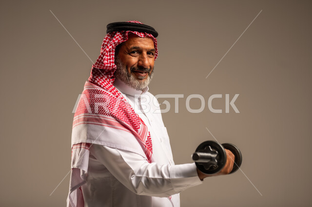 تمارين رفع الأثقال (دمبلز)، ممارسة التمارين الرياضية مع الدمبل، بورتريه مقرب لرجل كبير السن عربي خليجي سعودي مبتسم يرتدي الشماغ والثوب التقليدي ينظر الى الكاميرا يقوم برفع الأوزان الحديدية الثقيلة، المحافظة على الصحة واللياقة الجسدية، خلفية بيج