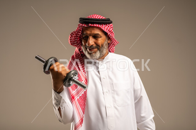 ممارسة التمارين الرياضية مع الدمبل، تمارين رفع الأثقال (دمبلز)، بورتريه مقرب لرجل كبير السن عربي خليجي سعودي مبتسم يرتدي الشماغ والثوب التقليدي يقوم برفع الأوزان الحديدية الثقيلة، المحافظة على الصحة واللياقة الجسدية، خلفية بيج