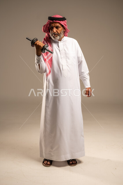 ممارسة التمارين الرياضية مع الدمبل، تمارين رفع الأثقال (دمبلز)، بورتريه لرجل كبير السن عربي خليجي سعودي مبتسم يرتدي الشماغ والثوب التقليدي يقوم برفع الأوزان الحديدية الثقيلة، المحافظة على الصحة واللياقة الجسدية، صورة الجسم بالطول الكامل، خلفية بيج