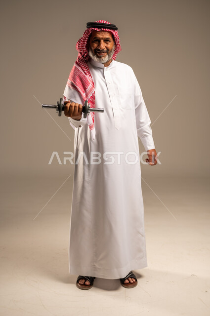 ممارسة التمارين الرياضية مع الدمبل، تمارين رفع الأثقال (دمبلز)، بورتريه لرجل كبير السن عربي خليجي سعودي مبتسم يرتدي الشماغ والثوب التقليدي ينظر الى الكاميرا يقوم برفع الأوزان الحديدية الثقيلة، المحافظة على اللياقة الجسدية، صورة الجسم بالطول الكامل، خلفية بيج