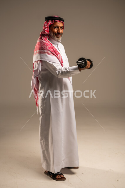 ممارسة التمارين الرياضية مع الدمبل، تمارين رفع الأثقال (دمبلز)، بورتريه لرجل كبير السن عربي خليجي سعودي مبتسم يرتدي الشماغ والثوب التقليدي ينظر الى الكاميرا يقوم برفع الأوزان الحديدية الثقيلة، المحافظة على اللياقة الجسدية، صورة الجسم بالطول الكامل، خلفية بيج
