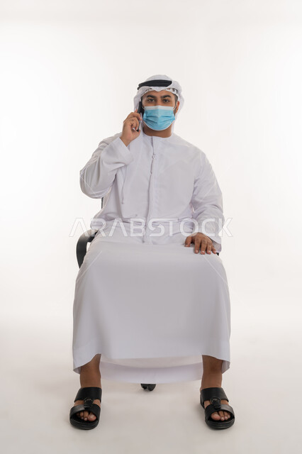 الالتزام باجراءات الحماية من فايروس كورونا, بورتريه لرجل عربي خليجي اماراتي يرتدي الغترة والكندورة البيضاء والكمامة الواقية يجلس على كرسي مريح وينظر الى الكاميرا، خلفية بيضاء