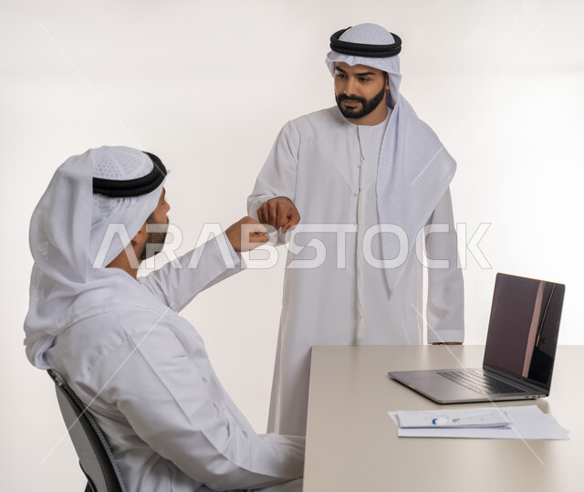 مفهوم التعاون بين الموظفين, التفاعل الجماعي لتحقيق أهداف الشركة, دمج التقنيات الحديثة المتطورة في الحياة العملية, بورتريه لموظفين عربيين خليجيين اماراتيين مبتسمين يرتديان الغترة والكندورة البيضاء يتصافحان بقبضة اليد, خلفية بيضاء