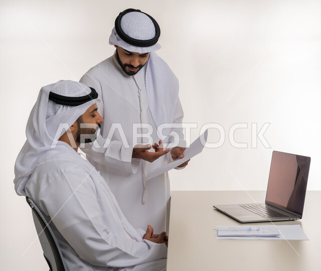 Collaborative management concept, close up side portrait of two Emirati Gulf Arab employees wearing white ghutra and kandura discussing work matters using papers and data, unity and team interaction to achieve company goals and solve problems, making new partnership deals, white background